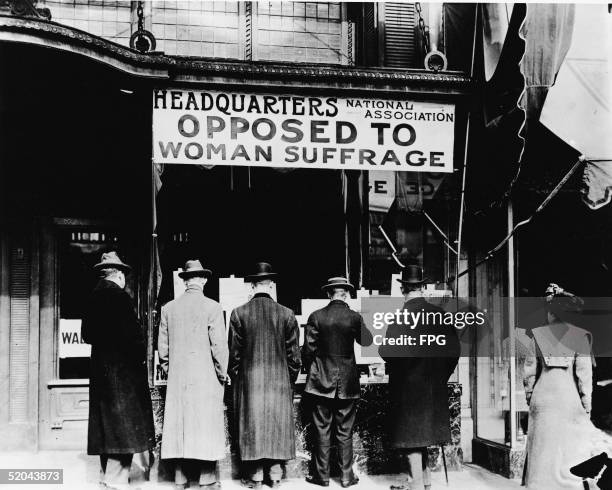 Men read literature posted in the window of the headquarters of the National Association Opposed to Woman Suffrage as woman stands off to the side,...