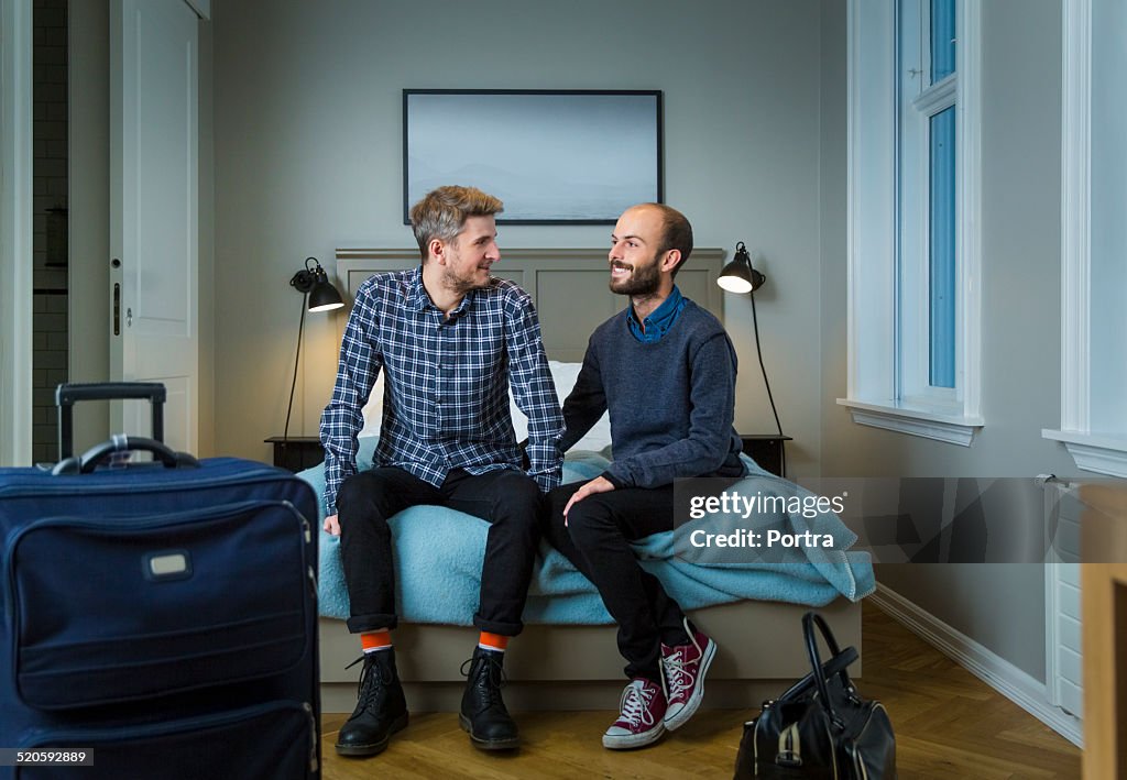 Happy gay men sitting on bed in hotel room