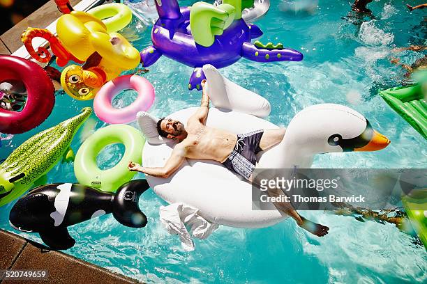 smiling man relaxing on inflatable swan in pool - abundance stock-fotos und bilder