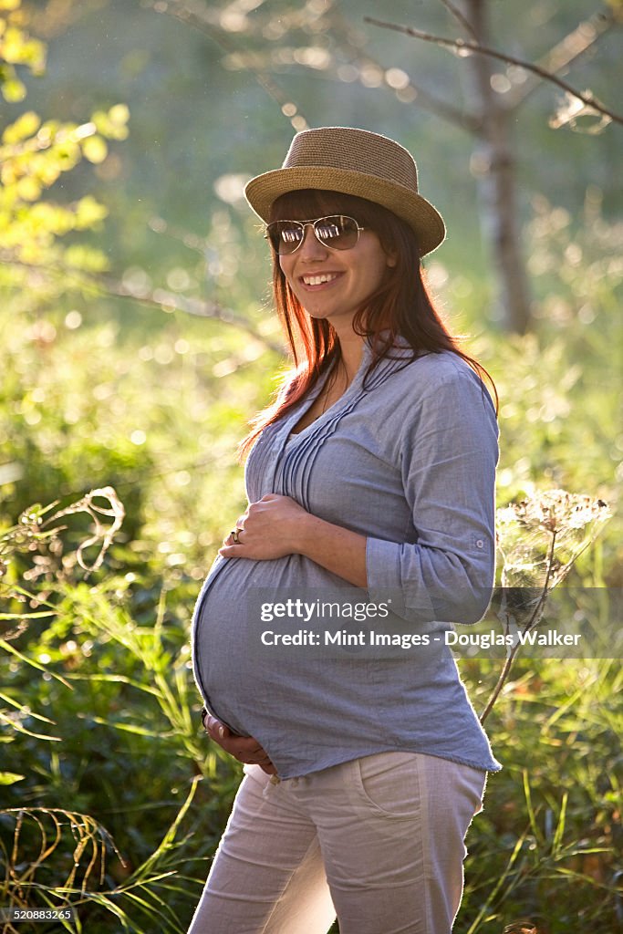 A pregnant woman in a hat and sunglasses, with her hand on her baby bump.