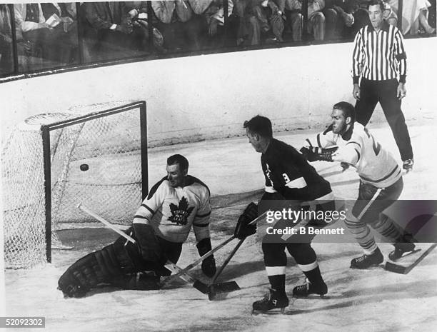 Gordie Howe of the Detroit Red Wings flips the puck into the net past goalie Johnny Bower of the Toronto Maple Leafs, while evading a check by Larry...