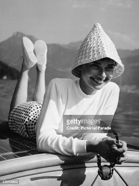 Portrait of Belgian-born American actress Audrey Hepburn wears a peculiar hat and lies on the back of a motorboat on a lake in Switzerland, early...