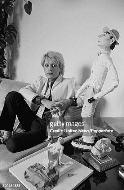 British musician and bass player with the group Japan, Mick Karn pictured sitting on a sofa with various artworks in London on 12th June 1980.