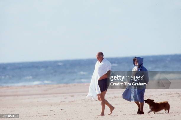 The Queen Mother With A Friend Walking Along The Beach With Her Pet Corgi. It Is A Windy Day So The Queen Mother Has Covered Her Hat With The Hood Of...