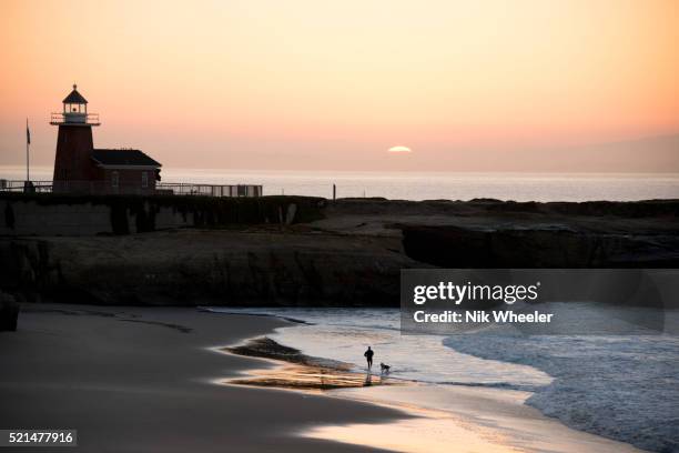 Woman walks her dog at sunrise on the sands of Lighthouse Field State Beach in front of the the former lighthouse, now Santa Cruz Surfing Museum, in...