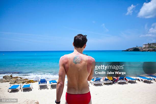 sunburned and tattooed man on beach using smart phone - sunburn tourist stock-fotos und bilder