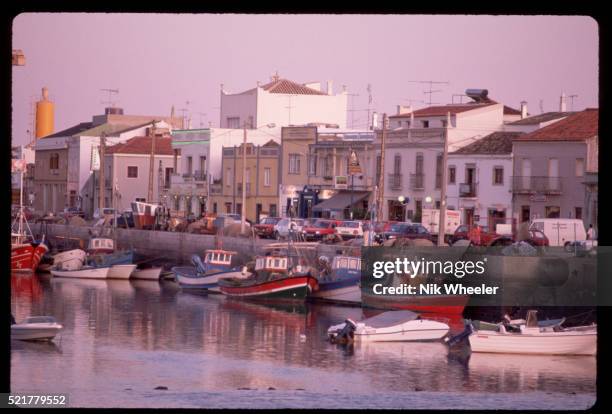 Colorful boats fill the harbor and businesses and cars line the street along the waterfront in Tavira, Algarve, Portugal.