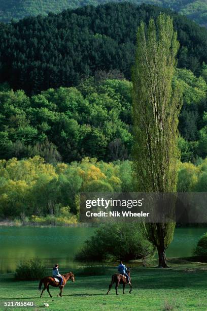 Two people ride horses along a river in the Abruzzo National Park.