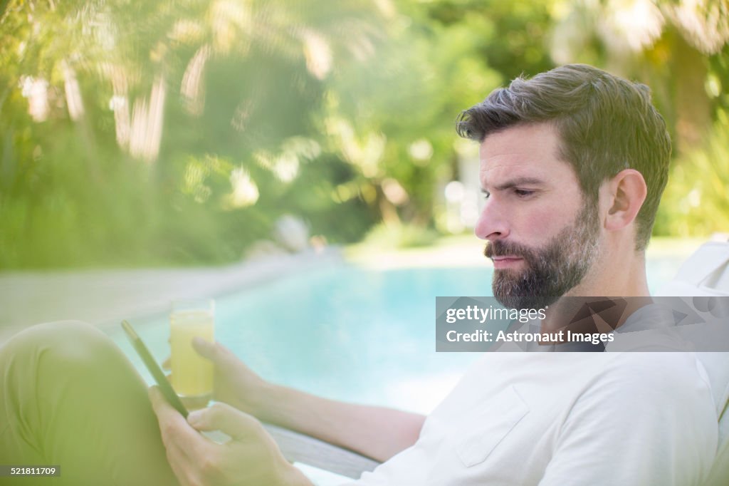 Man using digital tablet and drinking juice by swimming pool