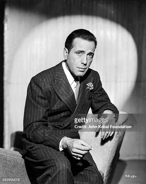 Posed portrait of actor Humphrey Bogart , wearing a suit and tie and smoking a cigarette, for Warner Bros Studios, 1946.