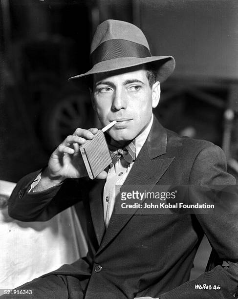 Posed portrait of actor Humphrey Bogart wearing a hat and bow tie with a pack of cigarettes, for Warner Bros Studios, 1938.