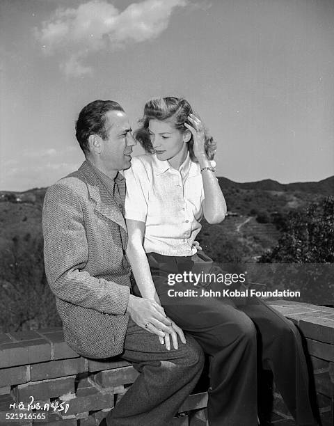 Actor Humphrey Bogart with his wife, actress Lauren Bacall , sitting together on a wall, for Warner Bros Studios, 1945.