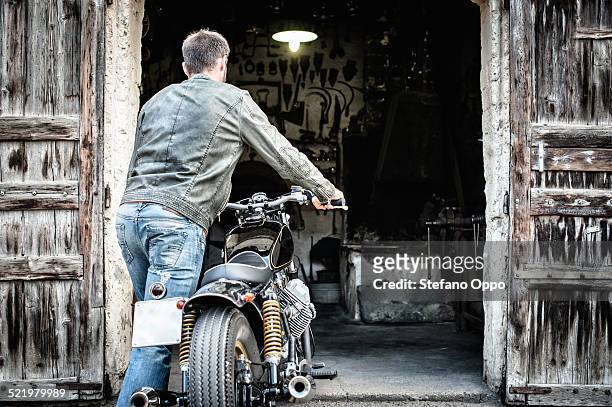 mid adult man pushing motorcycle into barn - man in denim jacket stock pictures, royalty-free photos & images