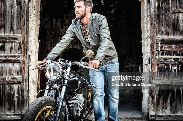 mid adult man pushing motorcycle out of barn - man in denim jacket stock pictures, royalty-free photos & images