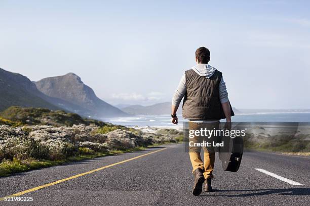 rear view of young man with guitar case walking on coastal road, cape town, western cape, south africa - vest fotografías e imágenes de stock