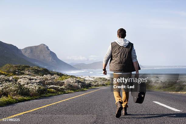 rear view of young man with guitar case walking on coastal road, cape town, western cape, south africa - étui à guitare photos et images de collection
