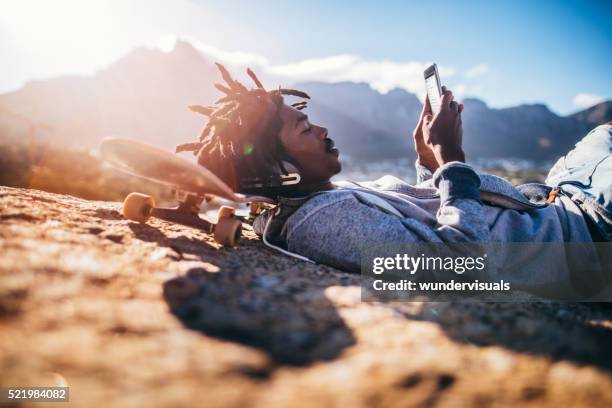 african american skater resting and looking at smartphone at sea - young skateboarder stock pictures, royalty-free photos & images