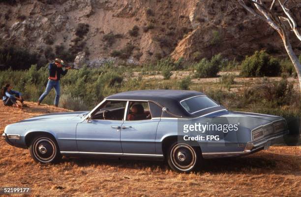 View of a 1969 Ford Thunderbird motor car parked outside while in teh background and women sitting on the grass watches a man in a Stetson and jeans...