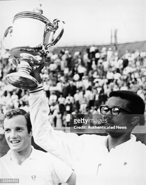 American tennis player Arthur Ashe holds up his trophy after winning the first ever US Open as his opponent Tom Okker of the Netherlands looks on and...