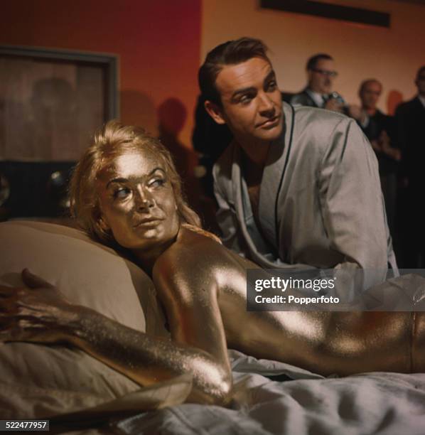 English actress Shirley Eaton pictured in character as Jill Masterson, lying on a bed covered in gold paint next to actor Sean Connery on the set of...