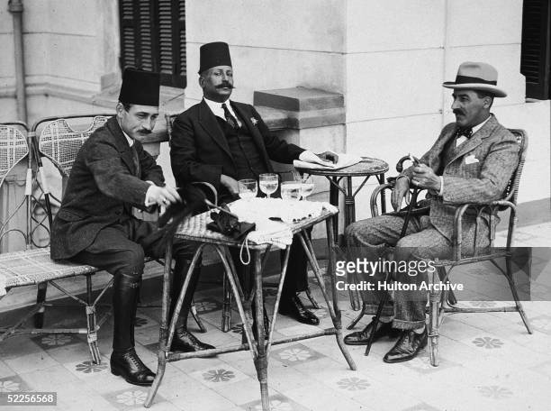 British archaeologist Howard Carter and two Egyptian officials sit on chairs at a table outside, Egypt, early 1920s.