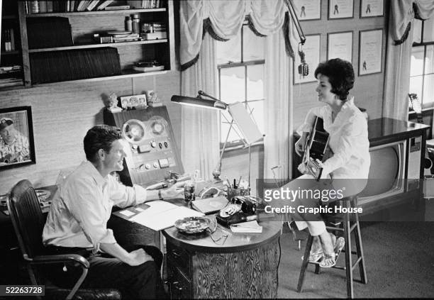 American country and western music singer and songwriter Harlan Howard sits at a desk next to a reel to reel recording device in an improvised...