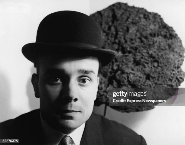Black and white portrait of French artist Yves Klein in a bowler hat as he stands in front of one of his Blue Sponge Sculptures, France, late 1950s....