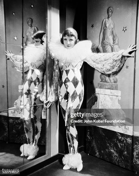 Portrait of actress Claudette Colbert standing in front of a mirror wearing a ruff around her neck and a sequined costume, as she appears in the...