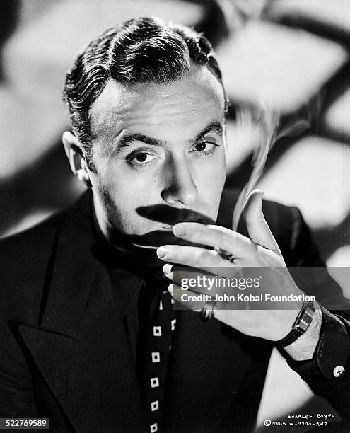 Portrait of actor Charles Boyer wearing a black shirt and tie and smoking a cigarette, for Columbia Pictures, 1938.