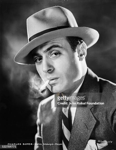 Headshot of actor Charles Boyer smoking a cigarette and wearing a hat, for MGM Studios, 1931.