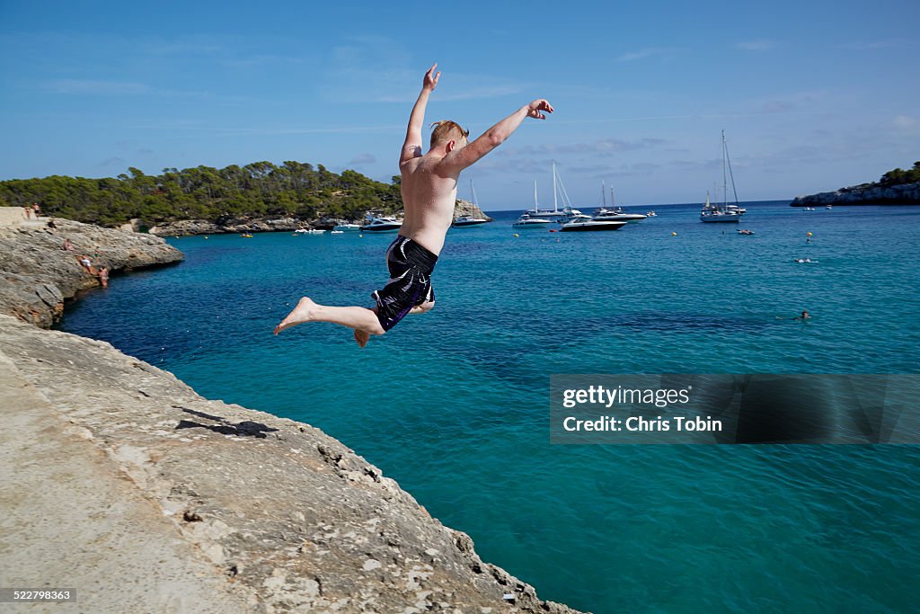 Young man jumping off rock into blue sea