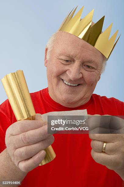 portrait of an aging man wearing a paper crown and looking at the joke inside his christmas cracker - coroa enfeites para a cabeça - fotografias e filmes do acervo