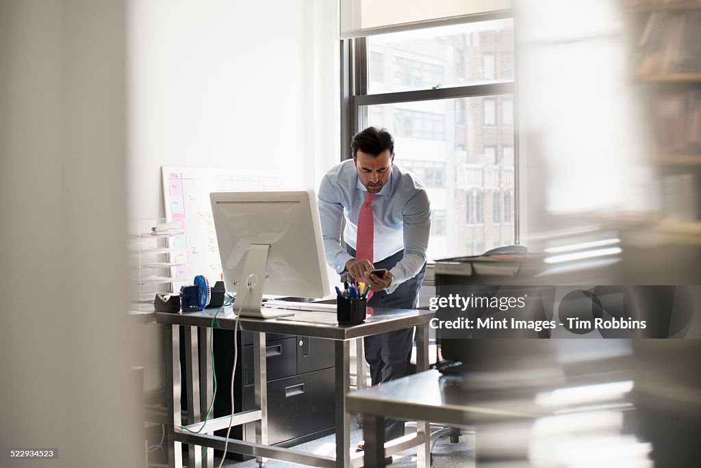 A man standing at his desk using his phone, dialling or texting.