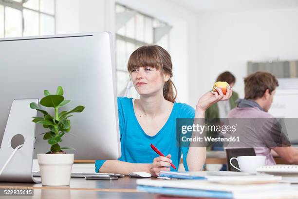 portrait of young woman eating an apple at her desk in a creative office - magazines on table stock pictures, royalty-free photos & images