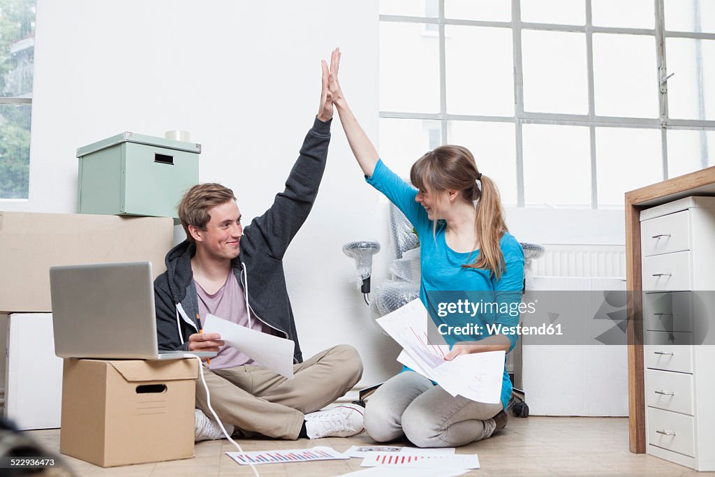 Two colleagues sitting on ground between cardboard boxes in an office giving high five