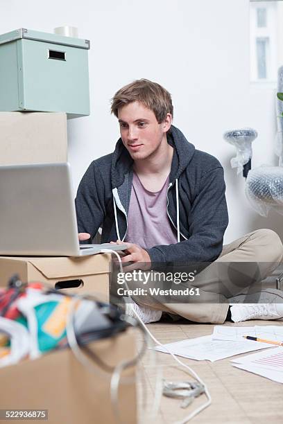 portrait of young man sitting on ground between cardboard boxes in an office using his notebook - makeshift office stock pictures, royalty-free photos & images