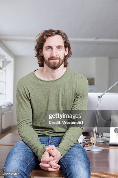 portrait of smiling young man sitting on a desk in an office - münchen immobilie büro stock-fotos und bilder