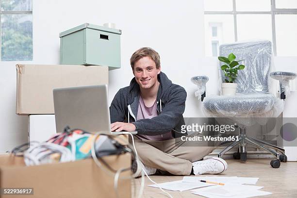 portrait of young man sitting on ground between cardboard boxes in an office using his notebook - makeshift office stock pictures, royalty-free photos & images