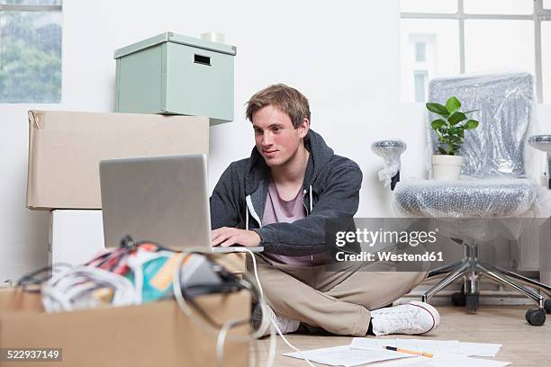 portrait of young man sitting on ground between cardboard boxes in an office using his notebook - makeshift stock pictures, royalty-free photos & images