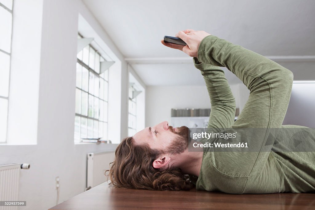 Young man lying on a table in an office taking a selfie with his smartphone