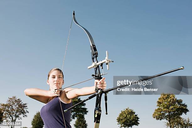 young woman aiming a bow and arrow - bogenschießen stock-fotos und bilder