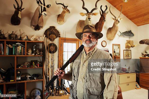 senior man with rifle standing in front of wall with various trophies - hunting stock pictures, royalty-free photos & images