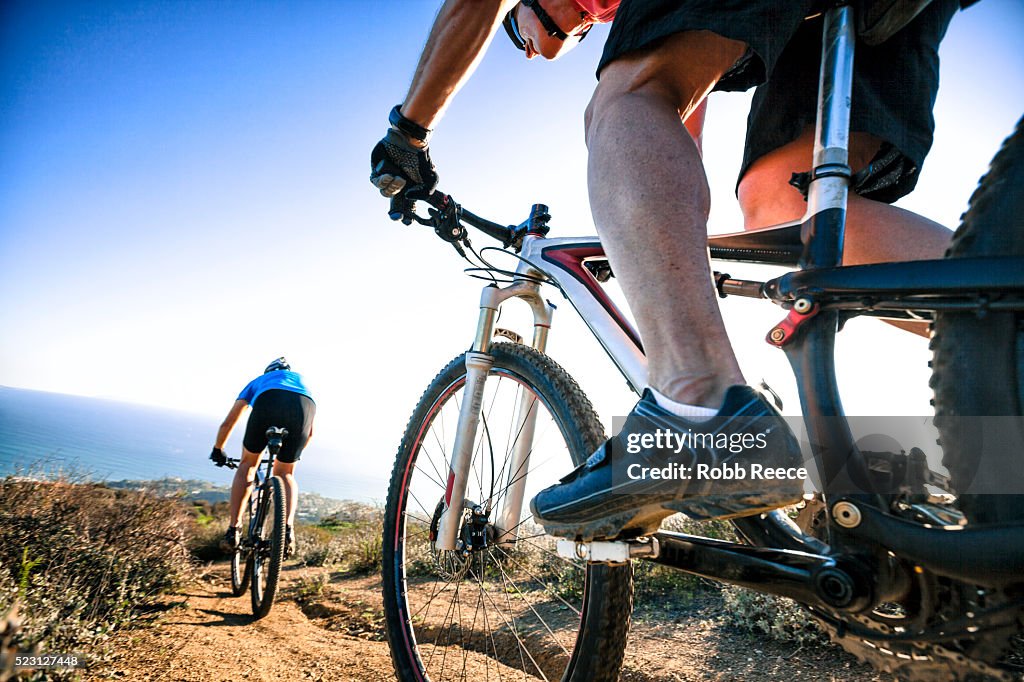 Two adult men riding mountain bikes on a steep, hilly trail above the ocean
