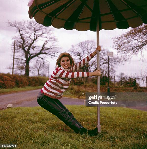 English actress Susan Hampshire posed wearing a red and white striped jumper and leather trousers whilst holding a large patio umbrella in 1964.