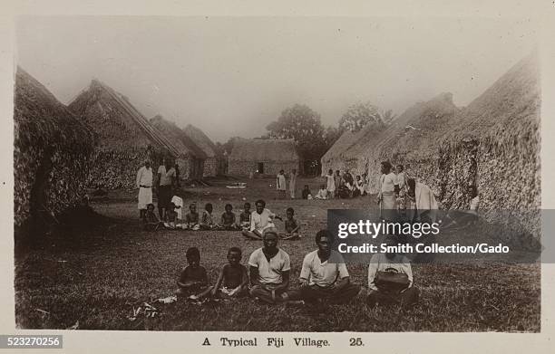 Postcard marked A Typical Fiji Village, showing a village lined with large huts, and several people walking and sitting on the ground, 1913. From the...