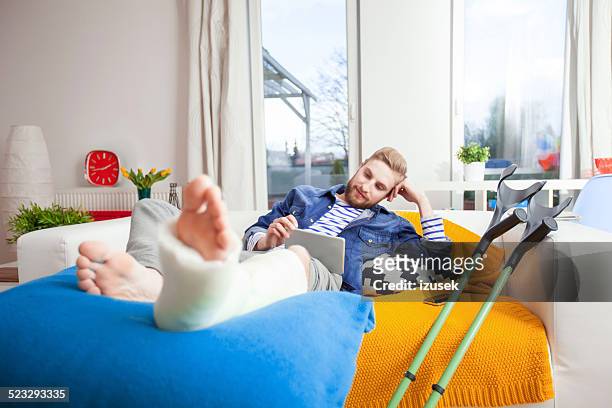 hombre joven con pierna fracturada usando tableta digital en su hogar - pierna fracturada fotografías e imágenes de stock