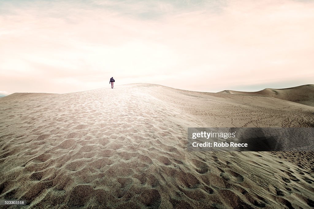 A man with a backpack hikes up a large sand dune in Death Valley, CA