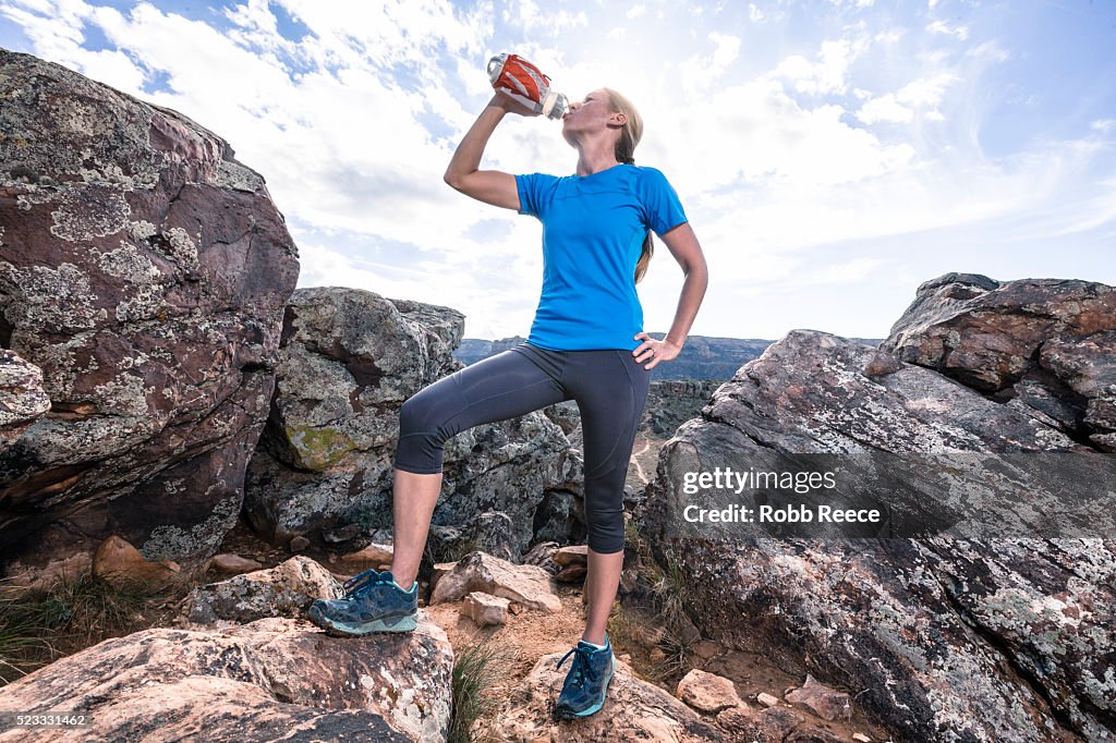 A woman trail runner on a rocky trail drinking from a water bottle