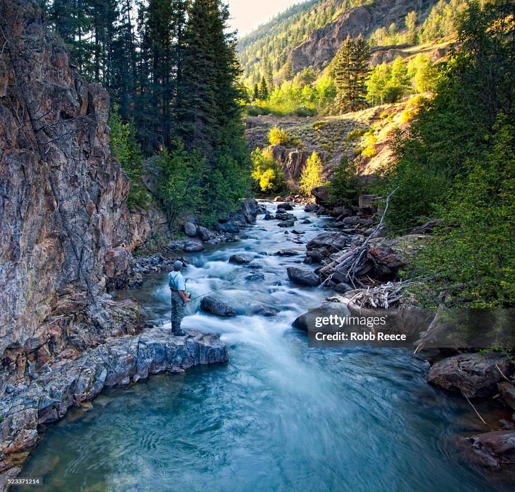 A male fly fisherman stands on a rock near a Colorado stream