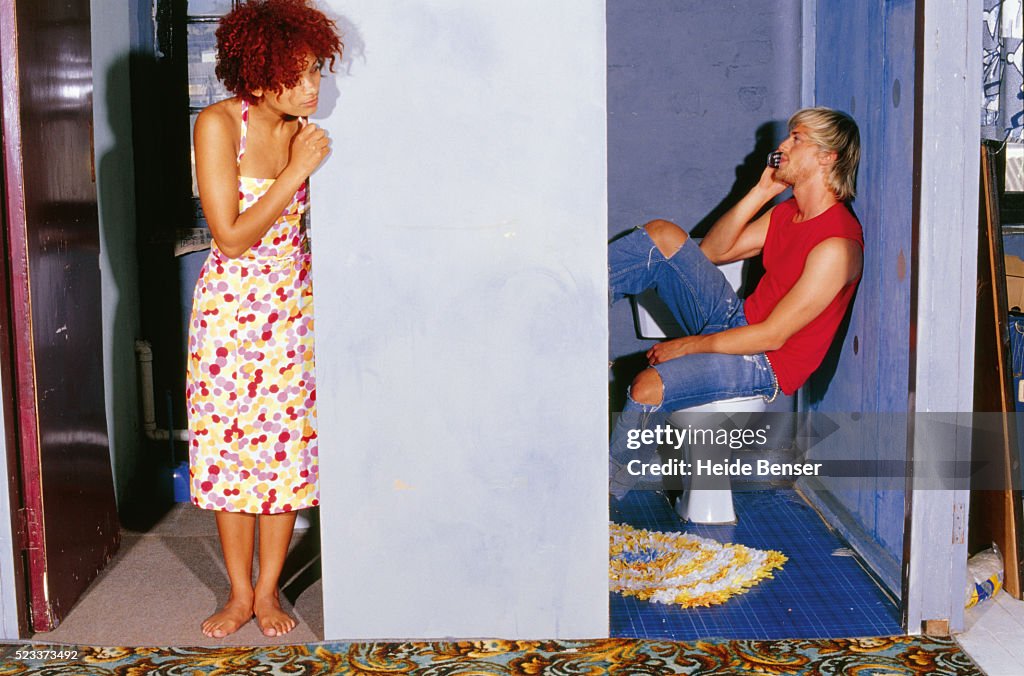 Young man on toilet using mobile phone, woman looking around the corner